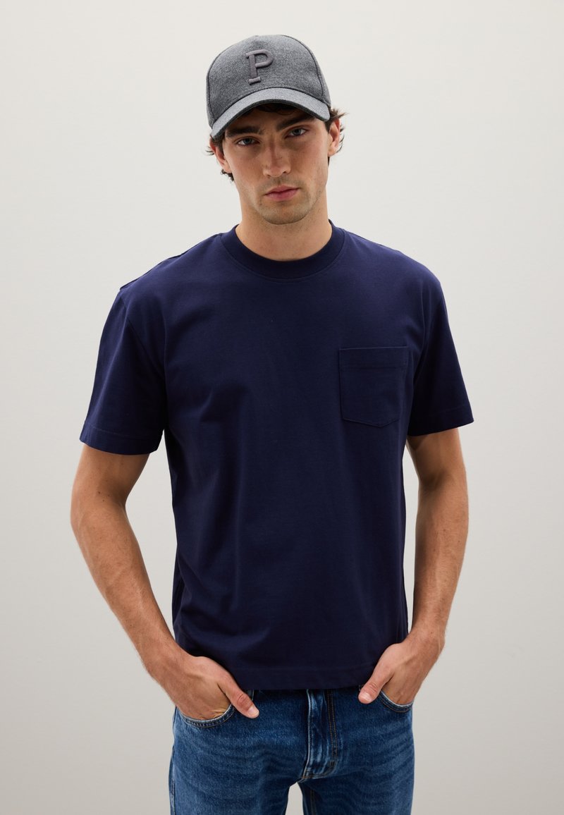 T-shirt en coton bleu marine avec un col rond et des manches courtes, doté d'une poche poitrine. Porté avec un jean en denim bleu clair et une casquette grise.