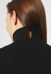 Donna con i capelli castani lisci che indossa un maglione nero a coste con un colletto alto e spaccato, vista da dietro e leggermente di lato.