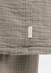 Nærbillede af beige, struktureret stof med synlig syning og en lille hvid mærkat med teksten "Originals" i kursiv skrift.