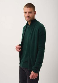 Tmavě zelený cardigan na zip vyrobený z jemného materiálu, s vysokým límcem a žebrovanými manžetami. Cardigan je nasazený přes odpovídající košili.