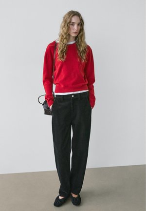 Roter Strickpullover, weißes Rundhals-Unterziehshirt, schwarze, weit geschnittene Hose aus strukturiertem Stoff und schwarze Slip-On-Schuhe. Kleine Handtasche mit Träger.