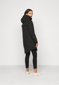 Abrigo largo negro con capucha, con una textura suave y una ligera forma en A, llevado sobre pantalones skinny negros y zapatillas de color claro.