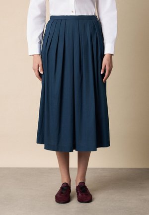 Donna che indossa una camicia bianca a maniche lunghe, gonna midi plissettata blu navy e scarpe bordeaux stringate, in piedi davanti a uno sfondo beige.