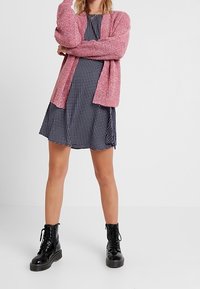 Cardigan lavorato a maglia rosa su un vestito nero a pois bianchi. Stivali neri con lacci chunky. Tessuto liscio con texture a contrasto.