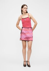 Νεαρή γυναίκα με ίσια καστανά μαλλιά που φοράει γυαλιά, ροζ κορυφή τύπου καμιζόλ, ροζ και μοβ φούστα τύπου περιτύλιγμα και μαύρα παπούτσια με τακούνια, στέκεται με τα χέρια πίσω.