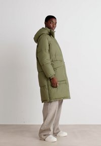 Olive green puffer coat with a hood, featuring a quilted design, side pockets, and elastic cuffs. Worn over light beige pants and white shoes.