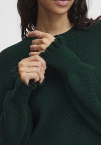 Dark green textured sweater with ribbed cuffs, featuring frilled edges. Model's hands show multiple thin silver rings.