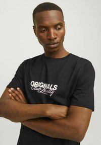 Camiseta de algodón negra con texto en blanco y un degradado rosa en negrita "ORIGINALS Club Ninety" en el pecho. Mangas cortas, diseño minimalista.