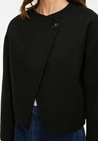 Giacca nera asimmetrica con bottoni, indossata sopra jeans di denim blu, che mostra la parte superiore del torso e il collo della persona.