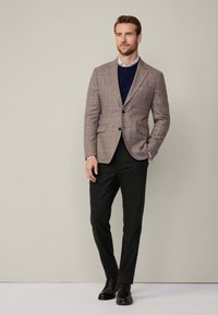 Hnědý kostkovaný blazer přes námořnický svetr, doplněný černými kalhotami a černými botami. Mezi významné detaily patří strukturované ramena a kapsy.