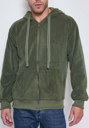 Homme portant un sweat à capuche zippé vert olive avec poches avant et cordons rayés, associé à un jean bleu, debout devant un fond blanc.