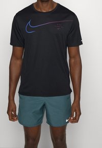 Svart Nike-t-shirt med korta ärmar, med en blå och lila swoosh-logotyp och text. Kombinerad med turkosa shorts och vita logodetaljer.
