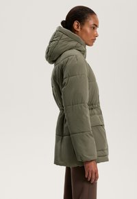 Olíva zöld pufi dzseki kapucnival, steppelt mintázattal, cinchelt derékkal és oldalsó zsebekkel. Sima textúra és puha felület.