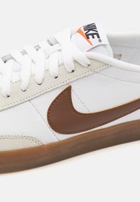 Sapatilhas brancas de couro com detalhe em camurça castanha, apresentando um swoosh da Nike cosido, atacadores brancos e uma sola de borracha texturizada.