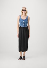 Jeans crop-top med knapper, sammen med en plissert svart maxi-skjørt. Modellen har på seg svarte åpen-toed mules og solbriller.