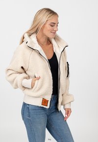 Kremowa polarowa kurtka z zapięciem na zamek, kapturem i czarnymi akcentami. Posiada boczne kieszenie oraz naszywkę z logo na dolnym brzegu.