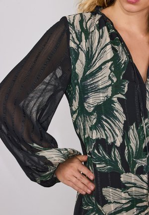 Donna indossa una camicetta nera trasparente con grande stampa floreale verde e beige e righe verticali strutturate, una mano appoggiata sul fianco.