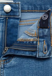 Sinisiä farkkuja, joissa on näkyvä metallivetoketju, hopeinen nappi ja ruskea neulonta. Sisäpuolella on kulumista ja tummansininen kangastunniste.