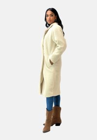 Abrigo de color crema de corte oversize con una tela suave y texturizada, dos bolsillos laterales y un ajuste relajado, que se lleva sobre unos vaqueros azules y botas marrones.