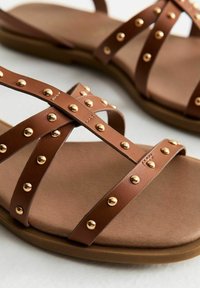 Bruna platta sandaler med korsade remmar prydda med gulddetaljer. Mjuk vadderad sula och en flexibel gummisula. Slät yta.