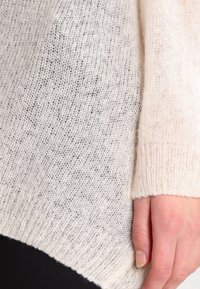 Pull en tricot beige clair avec une texture lâche et aérienne et un ourlet légèrement irrégulier. Le tissu présente de subtiles taches de tons plus foncés.
