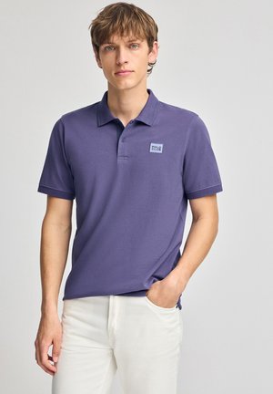 Camisa polo morada de algodón, con un cuello corto, tres botones y un pequeño logo cuadrado en el lado izquierdo del pecho. Combinada con pantalones blancos.