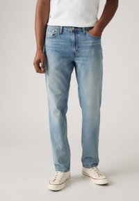 Calças jeans de ganga azul claro com corte direito, desbotamento leve, design de cinco bolsos e detalhes costurados, combinadas com ténis brancos.