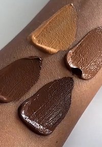 Fire farveprøver af foundation i forskellige nuancer - lys beige, medium tan og to dybe brune - påført en hudfarvet arm, der viser tekstur.