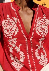 Röd broderad blus med vita blommönster, knappstängning och volangdetalj vid halsringningen, tillverkad av lättviktsmaterial.