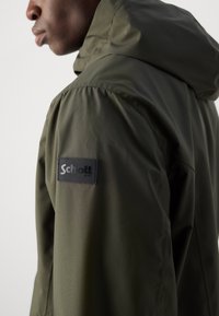 Olive green hooded jacket made of smooth, lightweight material. Features a black logo patch on the upper arm. Structured design, fitted silhouette.