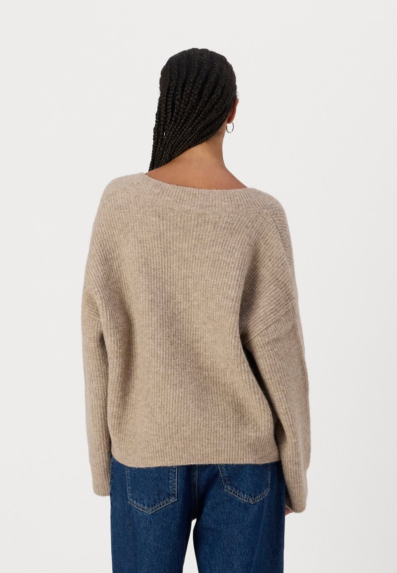 Beiger Strickpullover mit weitem Ausschnitt, lässiger Passform und gerippter Textur. Kombiniert mit blauen Jeans aus Denim, die minimale Nähdetails aufweisen.