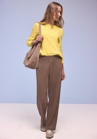 Jeune femme portant un pull jaune, un pantalon marron, des baskets beiges, avec un grand sac à bandoulière beige, debout devant un fond bleu pâle.