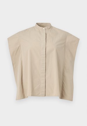 Naine seisab, kandes beeži varrukateta nööpidega tuba, valget asümmeetrist seelikut, pruune sandaale ja hoides väikest tumedat pruunist kotti.