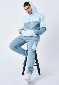 Sudadera azul claro con texto blanco y un bolsillo frontal, combinada con joggers a juego que tienen un bolsillo lateral con cremallera. Tejido suave, ajuste casual.