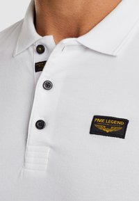 Kadar bijele polo majice s crnim gumbima i crno-žutim značkom PME Legend na prsima.