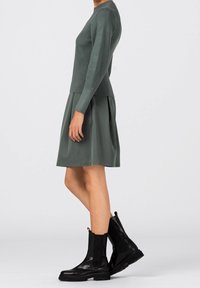 Ciemnozielony prążkowany sweter w połączeniu z dopasowaną rozkloszowaną spódnicą. Modelka nosi czarne botki z połyskującym wykończeniem i elastycznymi bokami.