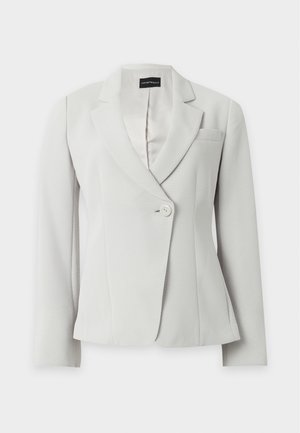Personne portant un costume gris clair et un haut blanc tient une grande pochette noire, debout devant un fond blanc uni.