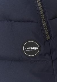 Granatowa kurtka pikowana z zamkiem błyskawicznym i okrągłą czarną naszywką z białym napisem. Materiał ma gładką, matową fakturę.