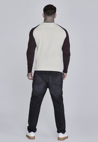 Langærmet skjorte med lys cremefarvet krop og mørkebrune ærmer, parret med sorte jeans og hvide sko. Har et sporty design og afslappet pasform.