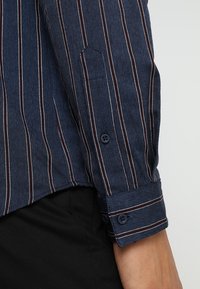 Mörkblå skjortärm med vita och röda vertikala ränder, knäppt manschett, bärs med svarta byxor.