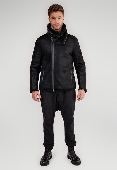 Uomo in giacca di shearling nera con zip asimmetrica, pantaloni larghi scuri e stivali neri con lacci, in piedi davanti a uno sfondo neutro.
