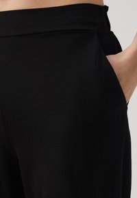 Černé kalhoty vyrobené z lehkého materiálu, s uvolněným střihem, bočními kapsami a hladkou texturou s jemným leskem.