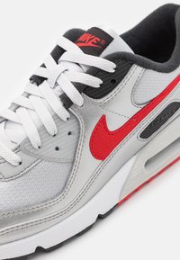 Baskets Nike Air Max blanches et grises avec logo swoosh rouge, lacets blancs, détails noirs et amorti Air Max visible dans la semelle.