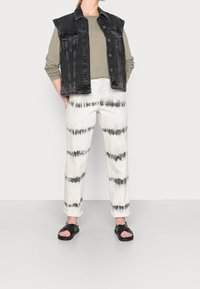 Person som bär en svart denimväst, olivgrön långärmad skjorta, vita tie-dye-byxor och svarta sandaler med öppen tå medan han står mot en enkel bakgrund.