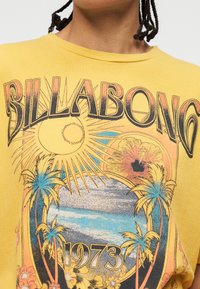 Κίτρινο πουκάμισο με κείμενο «Billabong 1973», έναν ήλιο, φοίνικες, κύματα ωκεανού και γραφικό σχέδιο με τροπικά λουλούδια.