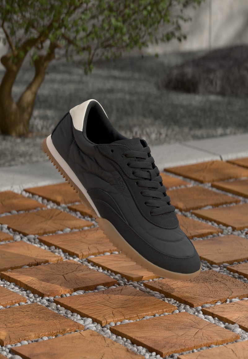 Scarpa da atletica nera con esterno in pelle liscia, accentata da un tallone bianco e suola in gomma testurizzata, posizionata su piastrelle di legno e ghiaia.