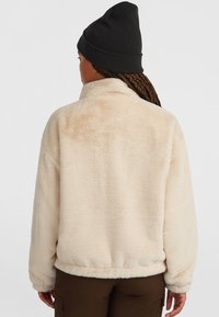 Fluffy beige vest met een hoge kraag, geribbelde zoom en lange mouwen. Gepaard met bruine broek en een zwarte gebreide muts. Zachte, textuurstof.