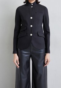 Chaqueta estructurada negra con un cuello alto, que cuenta con seis botones plateados, dos bolsillos con solapa y un corte entallado. Combinada con pantalones de cuero negros de pierna ancha.