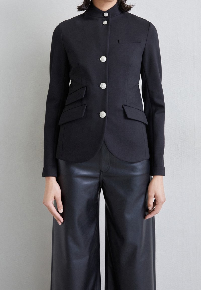 Chaqueta estructurada negra con un cuello alto, que cuenta con seis botones plateados, dos bolsillos con solapa y un corte entallado. Combinada con pantalones de cuero negros de pierna ancha.