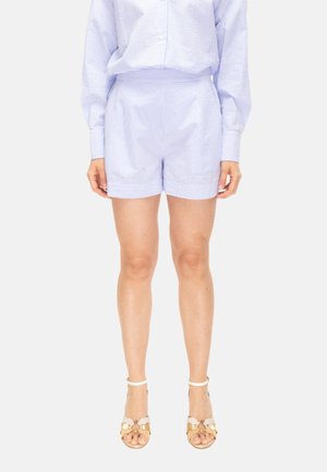 Femme portant un short rayé bleu clair et une chemise à manches longues assortie, avec des sandales à talons ouvertes beige et blanches.